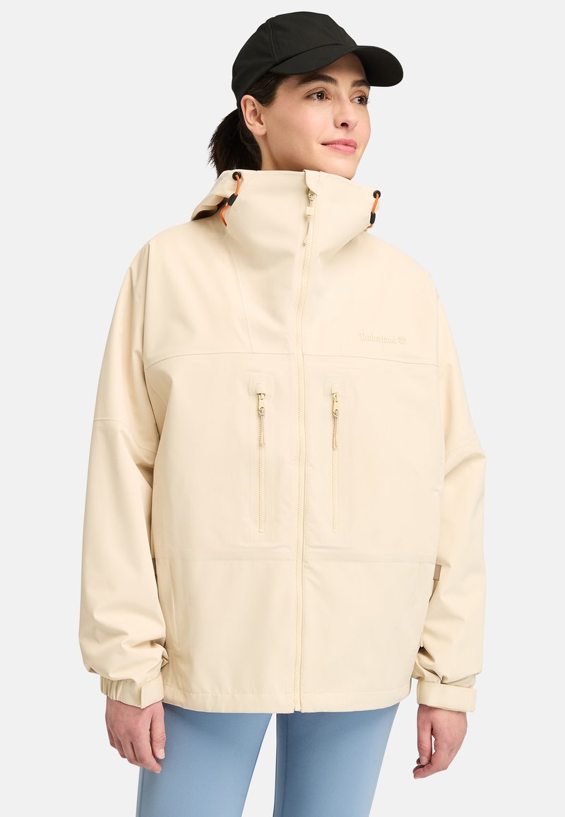 Lichtbeige waterdichte jas met een hoge kraag, twee ritszakken op de borst, verstelbare manchetten en oranje accenten. Zachte textuur en relaxte pasvorm.