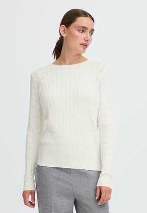 White knitted sweater with a textured cable pattern, round neckline, and long sleeves. Fitted at the waist, paired with gray trousers.
