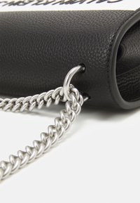 Černá texturovaná kožená taška s kovovým stříbrným řetízkovým popruhem, s hladkým, cylindrickým tvarem a viditelným embosovaným logem.