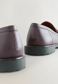 Paar dunkel bordeauxfarbener, strukturiertlederner Loafers mit dicken schwarzen Sohlen, von hinten auf hellem Untergrund fotografiert.