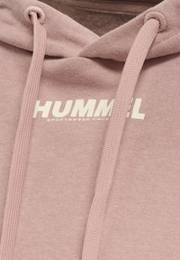 Gros plan d'un sweat à capuche rose avec cordons et texte blanc indiquant "HUMMEL SPORTSWEAR SINCE" sur le tissu.