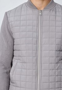 Veste bombardier matelassée grise avec un devant en tissu lisse, des détails côtelés aux manches, une fermeture éclair intégrale et des poches latérales.