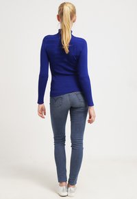 Blauer gerippter Rollkragenpullover mit langen Ärmeln, kombiniert mit dunklen Skinny Jeans und silbernen Schuhen, von hinten betrachtet.