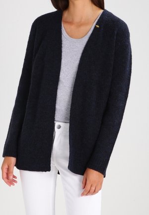 Nainen pukeutuneena tummansiniseen avoinna olevaan neuletakkiin, vaaleanharmaaseen pyöreäkauluksiseen t-paitaan ja valkoisiin housuihin, seisomassa yksiväristä taustaa vasten.