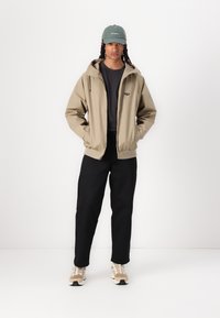 Tan zip-up jacket with a hood, black pants, and beige sneakers. The person is wearing a green cap, paired with a dark shirt underneath.