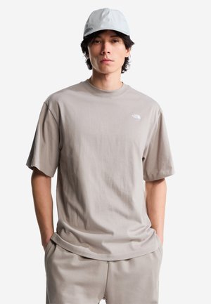 Joven con camiseta casual de color gris claro, pantalones a juego y gorra blanca, de pie con las manos en los bolsillos contra un fondo blanco.