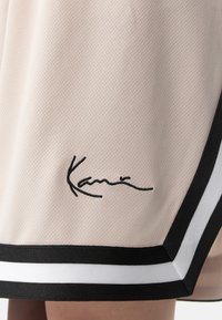Beige verkkoshortsit, joissa on musta-valkoinen raidallinen helma ja mustalla brodeerattu signeeraus, lisäävät huomattavaa yksityiskohtaisuutta muotoiluun.