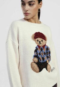 Maglione lavorato a maglia color crema con grafica di un orsacchiotto che indossa un cappello rosso, un gilet a rombi blu e pantaloni marroni. Collo e orlo a costine distintivi.