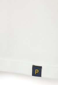 Tela blanca con una superficie texturizada, que presenta una pequeña etiqueta azul marino con una "P" amarilla en el dobladillo. Diseño limpio y detalles sencillos.