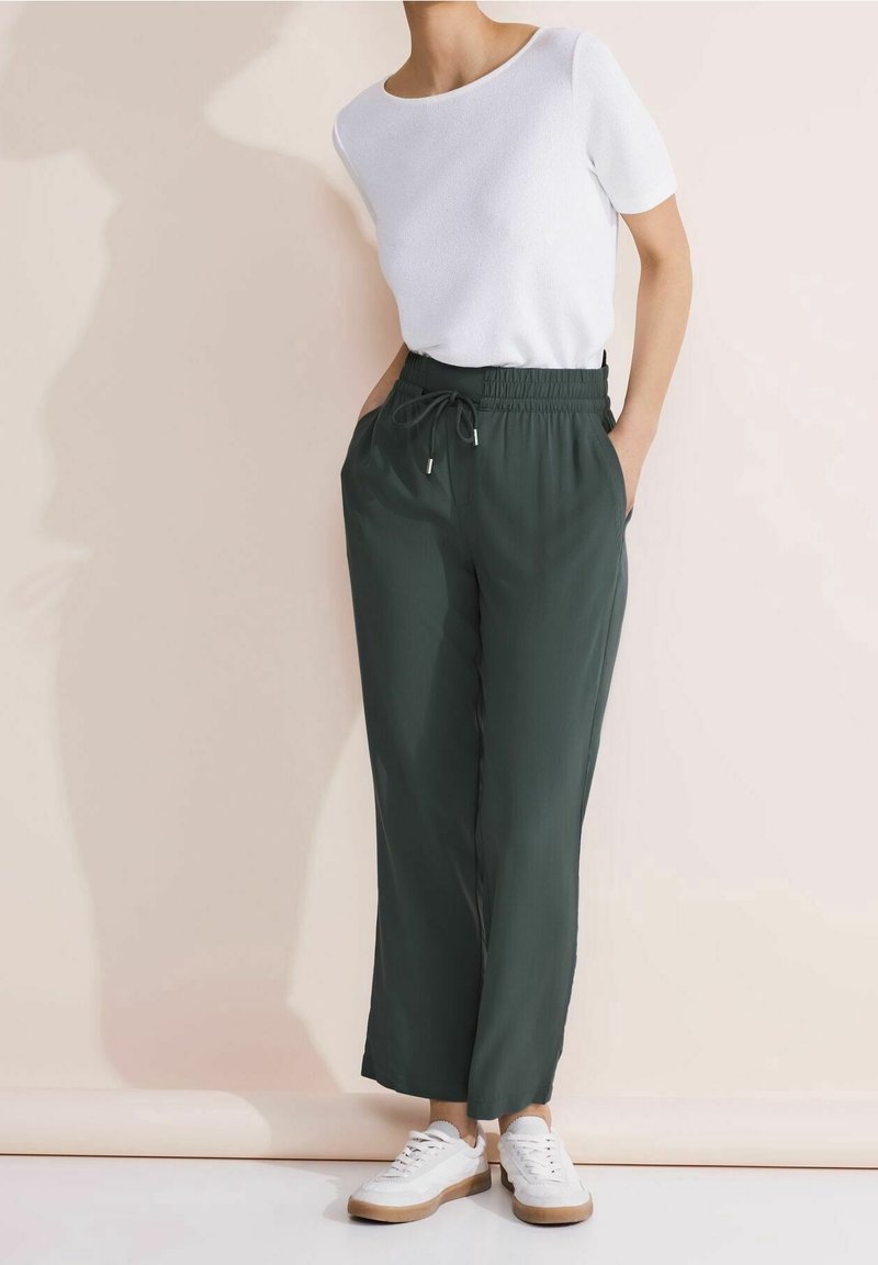 Personne portant un haut blanc à manches courtes, un pantalon vert foncé à cordon, et des baskets blanches, debout devant un fond beige clair.
