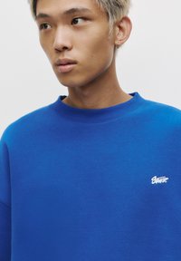 Sweatshirt bleu en tissu doux, avec un col rond côtelé et un petit logo blanc sur le côté gauche de la poitrine.