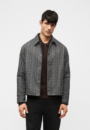 Άνδρας που φοράει γκρι καρό σακάκι πάνω από σκούρο καφέ πόλο και μαύρο παντελόνι, στέκεται μπροστά σε απλό ανοιχτό φόντο.