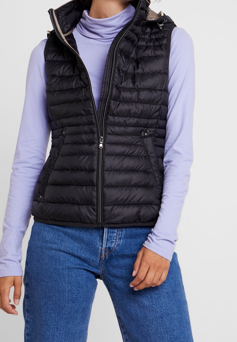 Kobieta ubrana w czarną pikowaną kamizelkę bez rękawów na jasnofioletowym golfie z długim rękawem i niebieskich dżinsach, stojąca w swobodnej pozie.