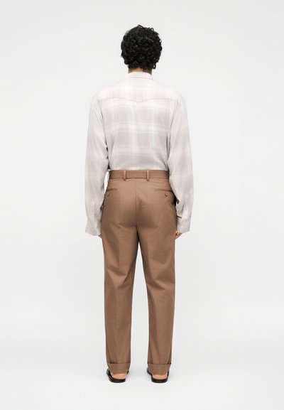 Άνδρας με σγουρά μαλλιά στέκεται με την πλάτη, φοράει ανοιχτόχρωμο καρό πουκάμισο που έχει μπει μέσα σε καφέ ραμμένο παντελόνι και μαύρα παπούτσια.