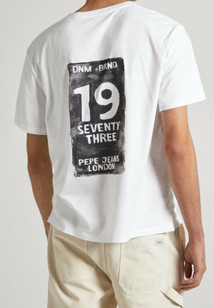 Mężczyzna w białej koszulce z czarnym prostokątnym nadrukiem z napisem "19 Seventy Three Pepe Jeans London" na plecach, widoczne beżowe spodnie.