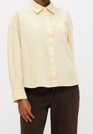 Persona que lleva una camisa de manga larga de color amarillo claro con rayas verticales y pantalones marrón oscuro.
