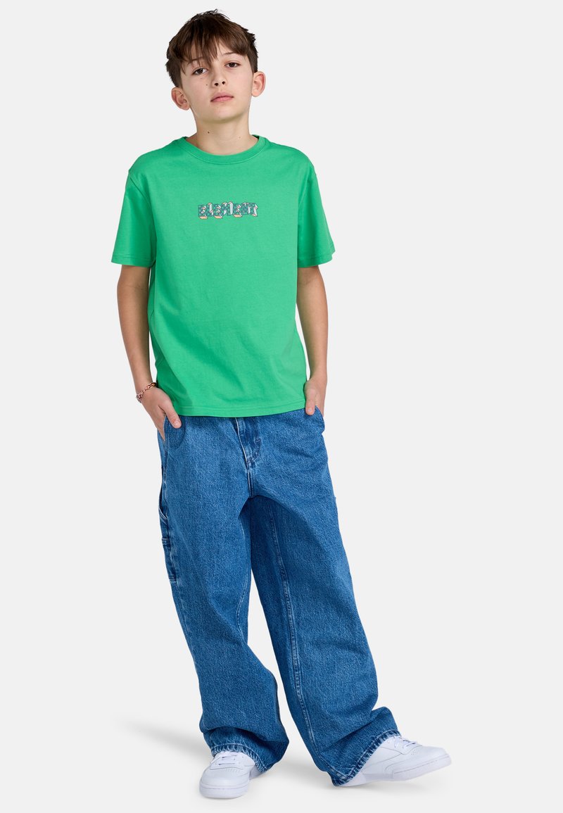 Zelené bavlněné tričko s grafickým textem na hrudi, kombinované s velkými modrými džíny. Bílí tenisky doplňují outfit.