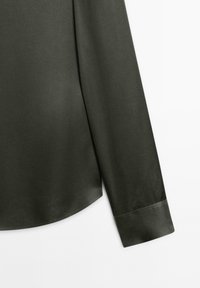 Tessuto della camicia verde scuro con una texture liscia, dotato di maniche con risvolto e orlo arrotondato, che sottolinea un design pulito e minimale.