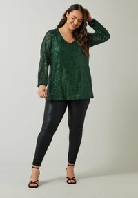 Grünes Paillettenoberteil mit V-Ausschnitt und langen Ärmeln, kombiniert mit schwarzen Lederleggings und schwarzen Riemchenheels. Glatte Textur.