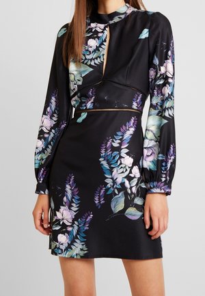 Kvinde iført en sort langærmet kjole med blå, lilla og hvide blomsterprint, med nøglehulshalsudskæring og en figursyet talje.