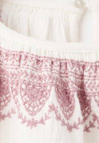 White fabric with light pink embroidery; features floral patterns and textured stitching along the neckline and bottom hem.