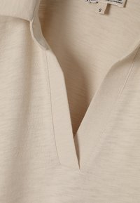 Béžové polo tričko s výstřihem do V, vyrobené z texturované látky. Obsahuje malou velikostní etiketu, což ukazuje na lehký a ležérní design.