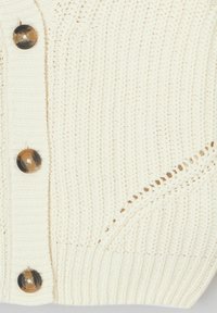 Gilet en tricot crème avec une texture côtelée, de grands boutons marron et un motif de mailles ouvertes diagonales sur le côté.
