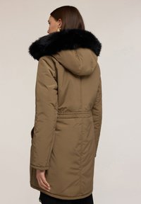 Cappotto imbottito marrone con colletto in pelliccia sintetica nera, cappuccio e vita con cintura. Presenta una texture trapuntata e tasche laterali per funzionalità.