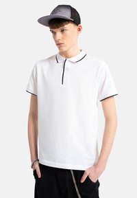 Biała koszulka polo z czarnym kołnierzykiem i wykończeniem rękawów, z trzema guzikami na plastrze. Model nosi również szaro-skalp z siateczkowym tyłem.