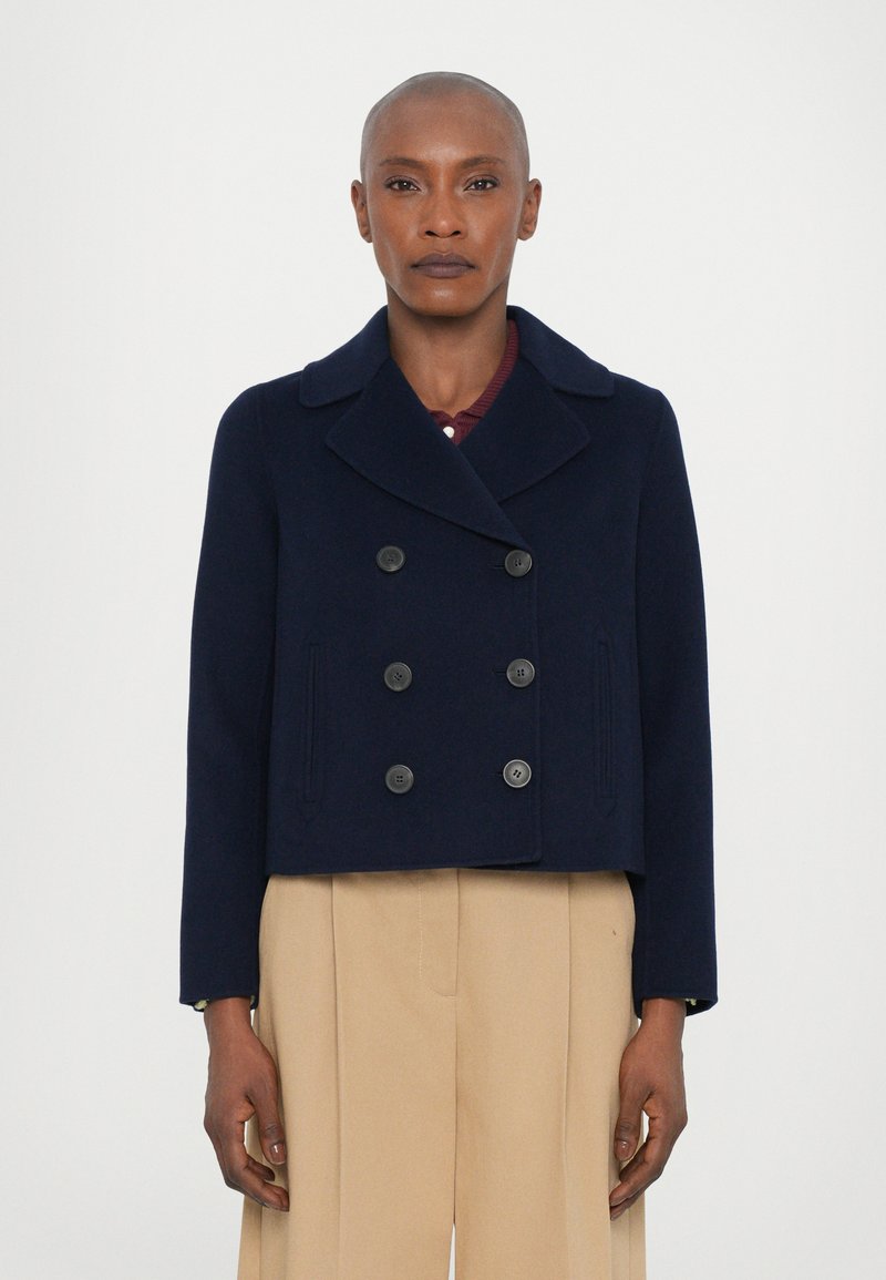 Giacca corta doppiopetto blu navy in lana, con ampia collaro e sei bottoni neri, abbinata a pantaloni beige.