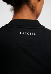 Svart polotröja med en texturerad krage, försed med en vit "LACOSTE"-logotyp på baksidan. Slät tyg med en figursydd design.
