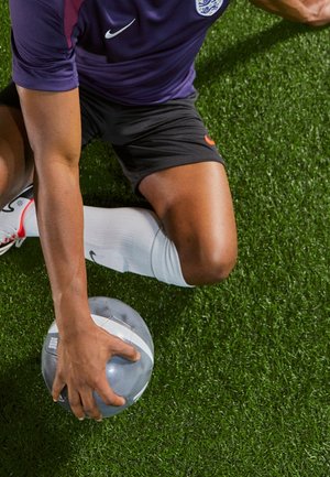 Camiseta deportiva de Nike en morado con un logo, pantalones cortos negros y calcetines blancos; modelo sentado sobre un césped artificial verde, sosteniendo un balón gris.