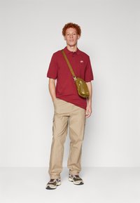 Maroon majica polo s kratkim rukavima, bež cargo hlače, maslinasto zelena torba preko ramena i šarena tenisica s teksturiranom potplatom.