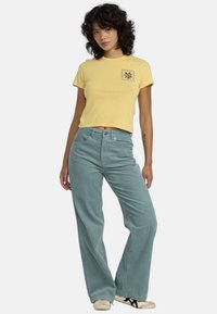 T-shirt jaune court avec une impression sur la poitrine, associé à un pantalon teal à jambes larges. Le modèle porte des baskets blanc cassé, affichant un style décontracté et relax.
