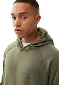 Olivgrön hoodie med snörning i huvan, texturerat tyg och broderad logo på bröstet. Har raglanärmar och en avslappnad passform.
