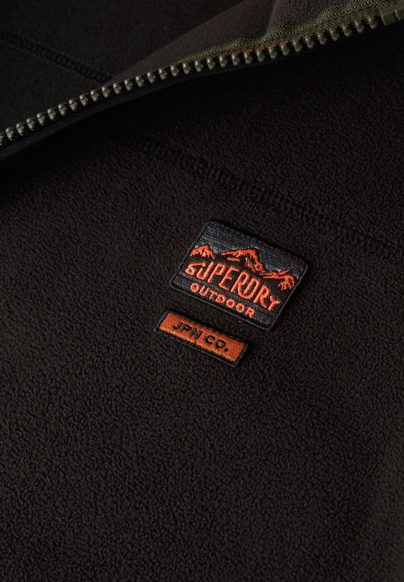 Veste en polaire noire avec un patch Superdry Outdoor et une petite étiquette JPN Co., dotée d'un col zippé de couleur contrastante.
