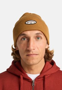 Bruine geribbelde gebreide beanie met een logo-opdruk, gedragen door een persoon met krullend haar, gecombineerd met een rode rits hoodie.