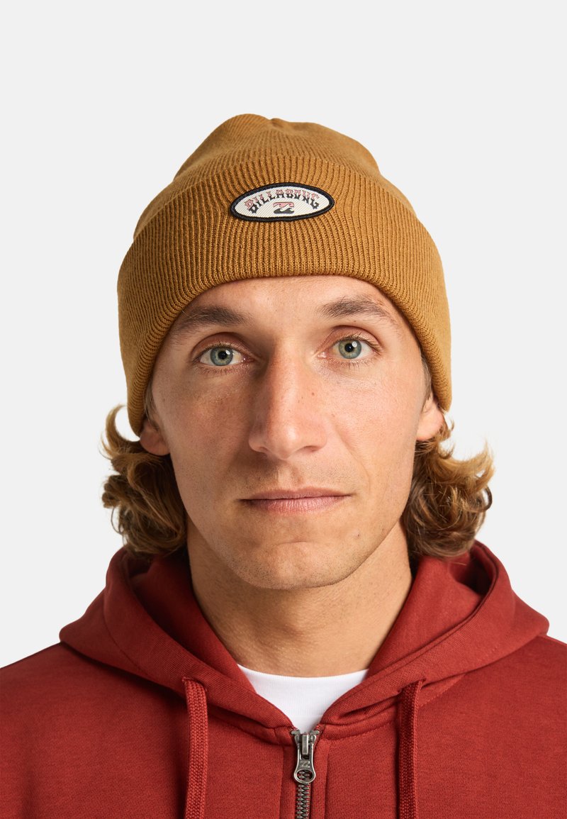 Bruine geribbelde gebreide beanie met een logo-opdruk, gedragen door een persoon met krullend haar, gecombineerd met een rode rits hoodie.