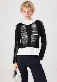 Schwarzer, verkürzter Pullover mit abgeschnittenem Design, der breite Ausschnitte und ausgefranste Kanten aufweist, über einem weißen Langarmshirt getragen.