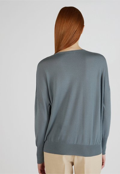 Grauer leichter Pullover mit lockerer Passform, geripptem Saum und langen Ärmeln. Glatte Textur mit subtiler Glanz. Hellbeige Hosen sind darunter sichtbar.