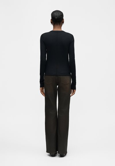 Γυναίκα με κοντά μαλλιά, φορώντας μαύρη μπλούζα με μακριά μανίκια και σκούρο παντελόνι φαρδύ μέχρι τον αστράγαλο, στέκεται με την πλάτη προς την κάμερα σε ένα μονόχρωμο φόντο.