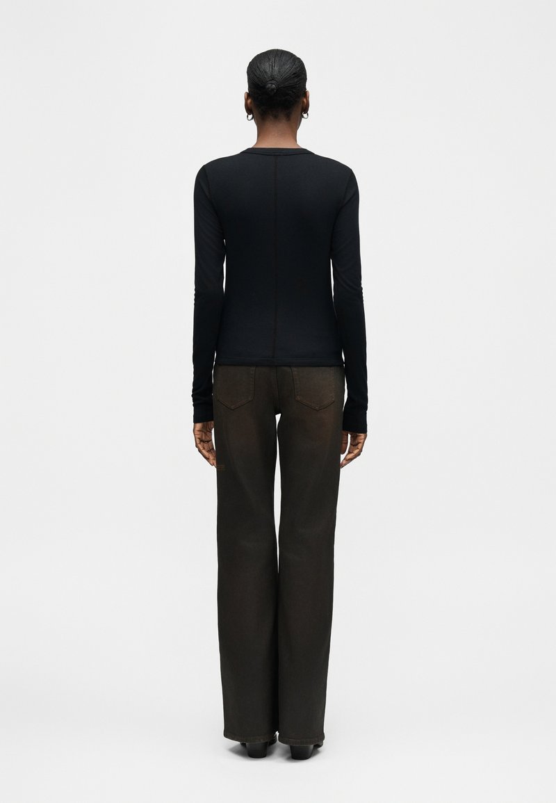 Woman with short hair in a black long-sleeve top and dark wide-leg pants standing with her back to the camera against a plain background.