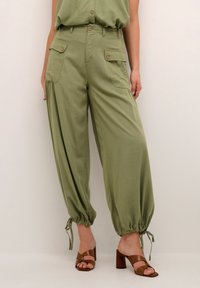 Olivově zelené, volné kalhoty s vysokým pasem, dvěma bočními kapsami a stahovacími manžetami. V kombinaci s hnědými sandály na blokovém podpatku.