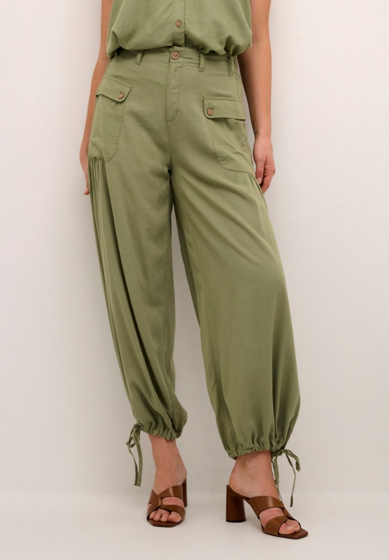 Olivově zelené, volné kalhoty s vysokým pasem, dvěma bočními kapsami a stahovacími manžetami. V kombinaci s hnědými sandály na blokovém podpatku.