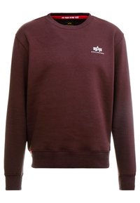 Mørkerød crewneck-sweatshirt med lange ærmer, hvid Alpha Industries-logo på brystet og et rødt inderhalsmærke med teksten "Remove Before Flight."