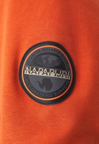 Tessuto arancione con lavorazione a maglia texturizzata. Presenta una toppa rotonda in gomma nera con la scritta "NAPAPIJRI" e una mappa dell'Antartide, accentuata da un bordo arancione.