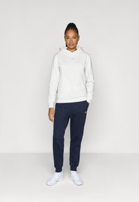 Hanorac alb cu glugă, având un logo violet, purtat cu pantaloni de trening albastru marin și adidași albi. Țesătură netedă, cu o croială lejeră.