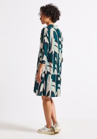 Abito stampato in teal, crema e navy con un design a strati. Presenta maniche a tre quarti e un colletto. Abbinato a scarpe da ginnastica bianche.