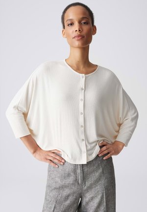 Femme les mains sur les hanches portant un chemisier blanc ample boutonné avec des manches trois-quarts et un pantalon gris texturé.