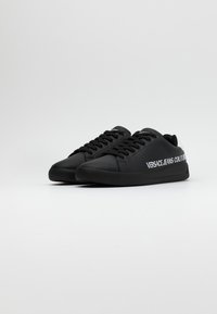 Svarta sneakers tillverkade av slät syntetiskt material. Funkar med rund tå, platt sula och snörningar. Texten "VERSACE JEANS COUTURE" på sidan.